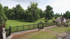 A photo of Revesby Estate. The image shows the estate grounds enclosed by a large iron fence and a regal gate. In the background well kept gardens contain a number of large trees and a house to the right.